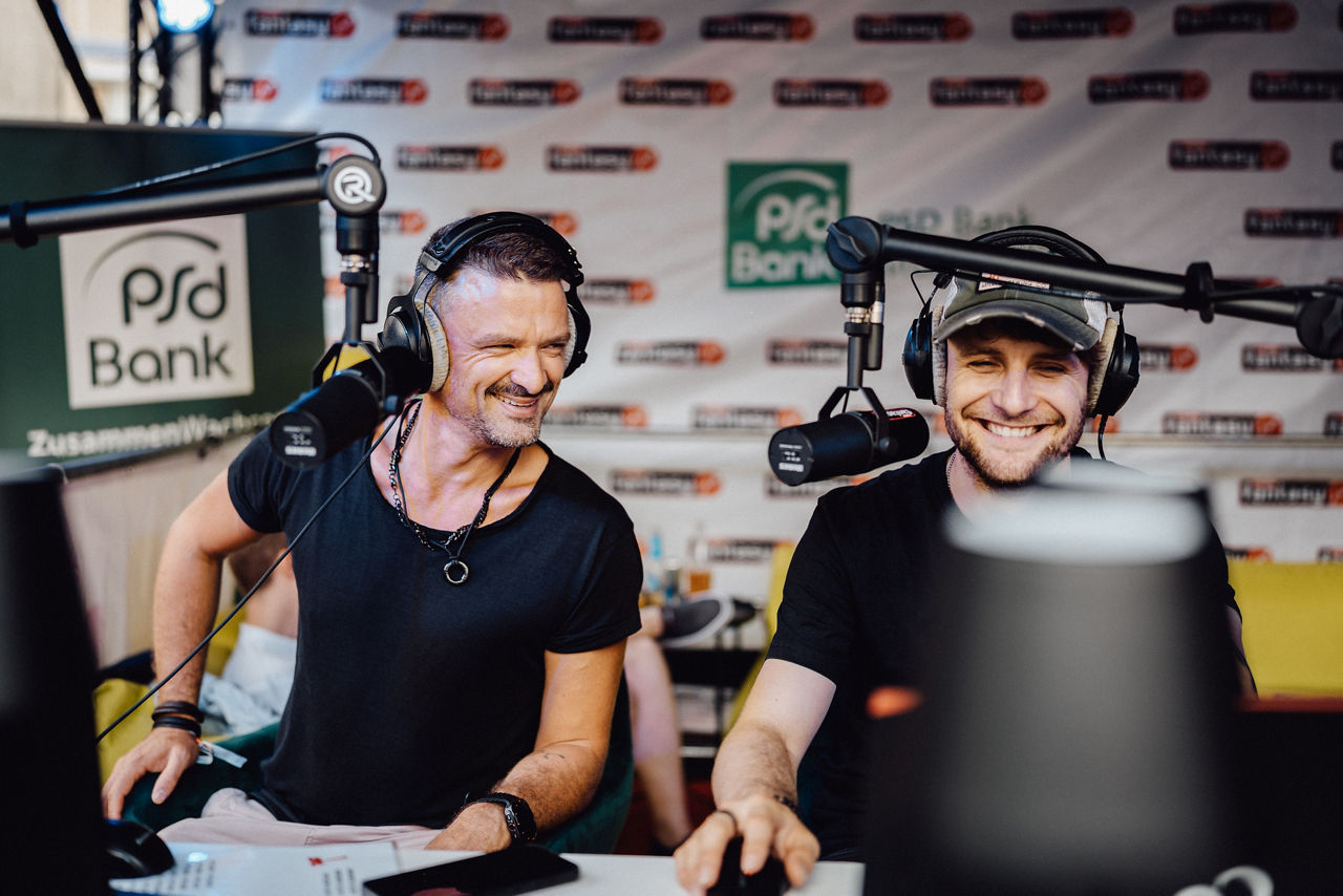 Im Vordergrund sitzen zwei Männer an einem Schreibtisch. Sie tragen Kopfhörer und jeder hat ein Mikrofon vor sich.  Beide lachen. Im Hintergrund ist eine Werbewand von Radio Fantasy und der PSD Bank München. 