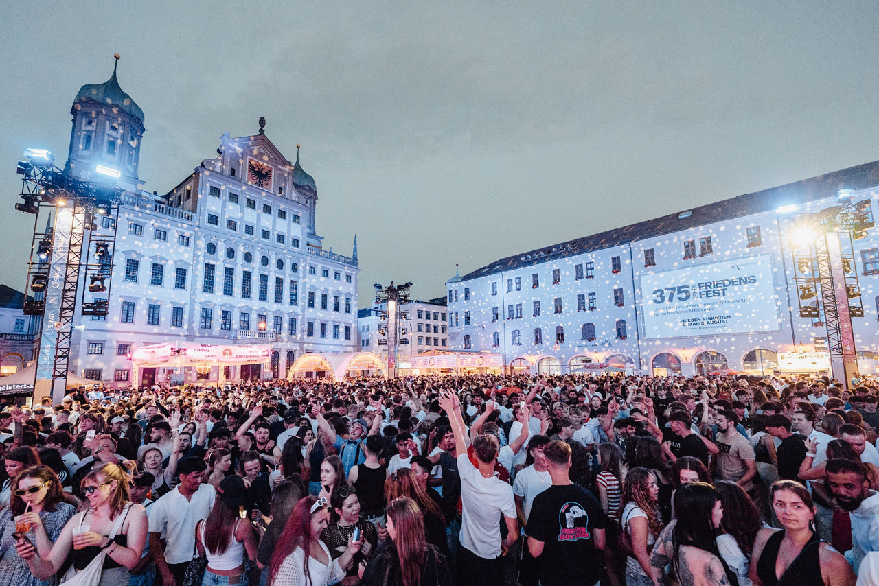 Im Hintergrund ist links das Augsburger Rathaus und rechts ein weiteres Gebäude. Beide werden in der Dämmerung beleuchtet. Im Vordergrund tanzen auf dem Platz davor eine Vielzahl an Menschen. 