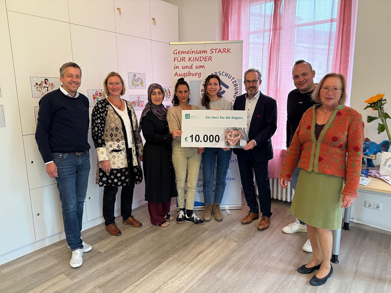 Acht Personen, drei Männer und fünf Frauen stehen nebeneinander. Übergeben wird ein Scheck in Höhe von 10.000 Euro. 