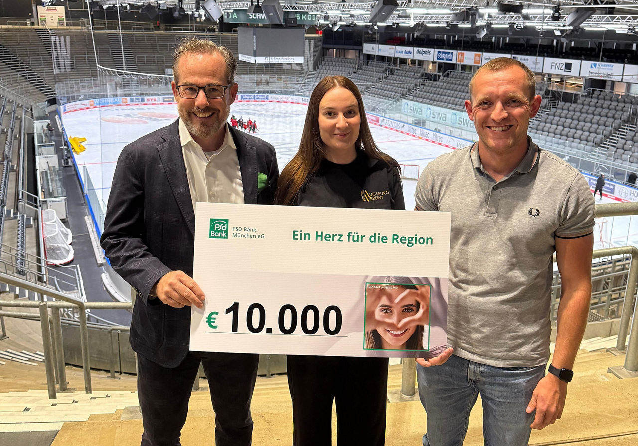 Drei Personen, zwei Männer und eine Frau, stehen in einem Eisstadion. Zusammen halten sie einen Scheck in Höhe von 10.000 Euro. 