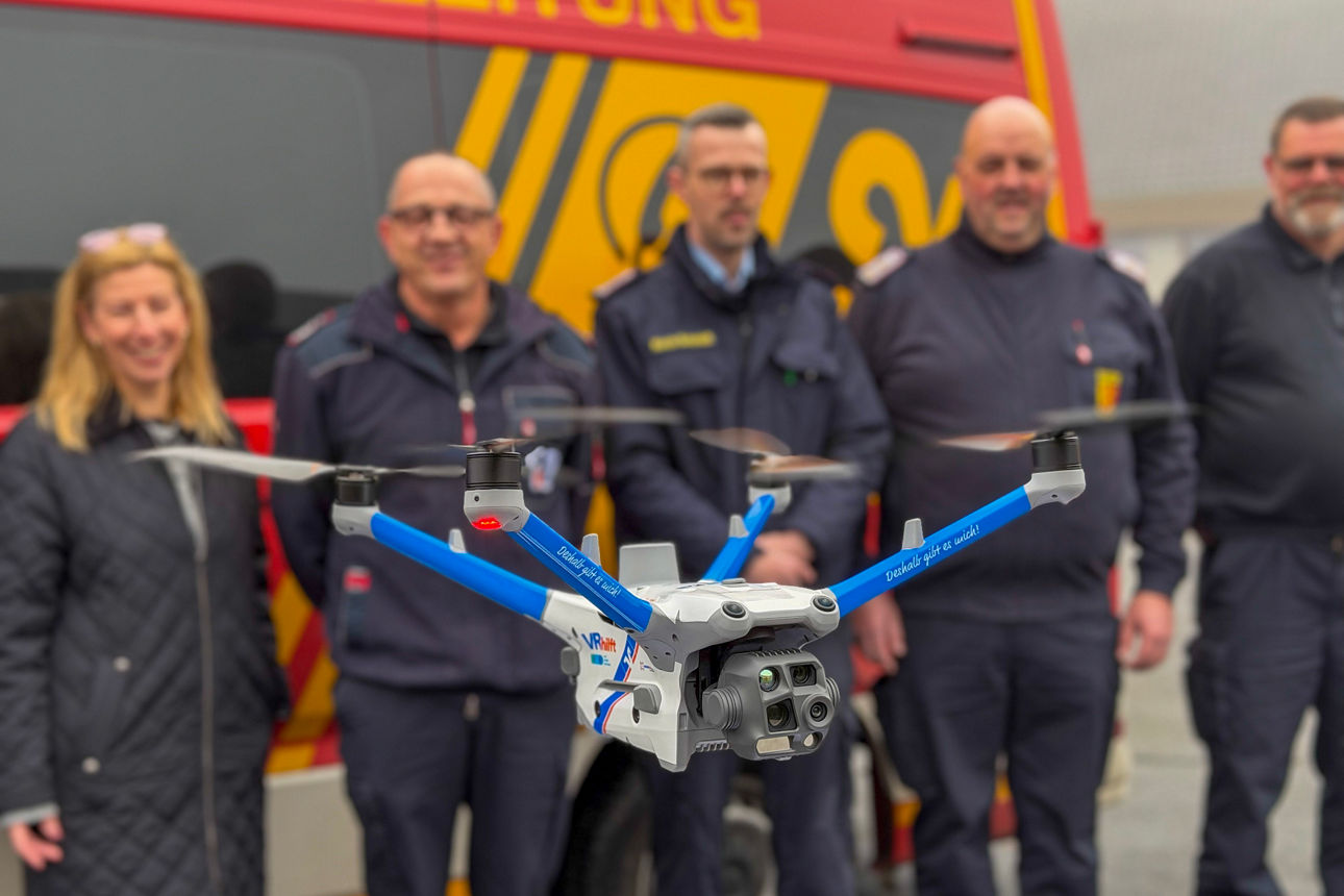 Im Vordergrund fliegt eine BOS-Drohne vom Typ M4TD. Im Hintergrund stehen Feuerwehrleute vor einem Rettungswagen.