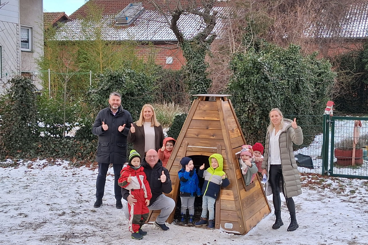 Ein Holz-Tippi-Haus in einem Garten besetzt mit Kindern und vier Erwachsenen, die sich freuen und die Daumen nach oben zeigen.