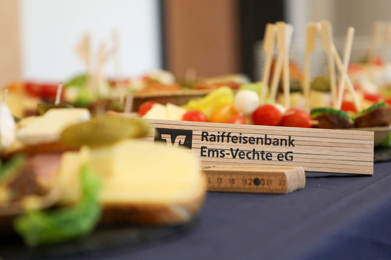Ein Zollstock mit Logo der Raiffeisenbank Ems-Vechte eG liegt dekorativ in einem Snack-Buffet