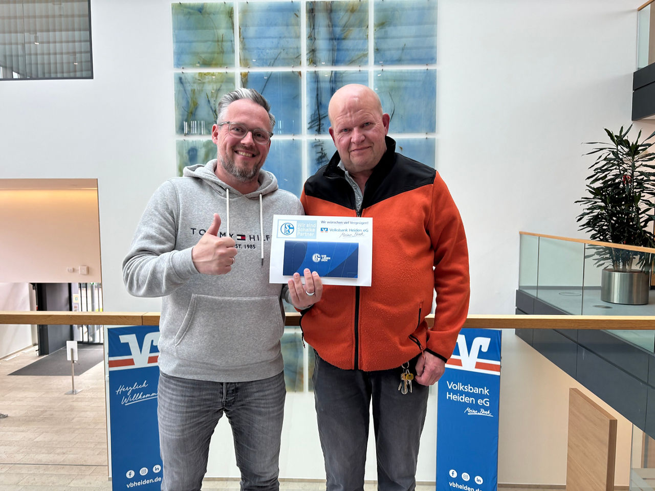 Zwei Männer stehen in der Galerie eines modernen Gebäudes und halten ein Zertifikat mit Markenlogo in der Hand.
