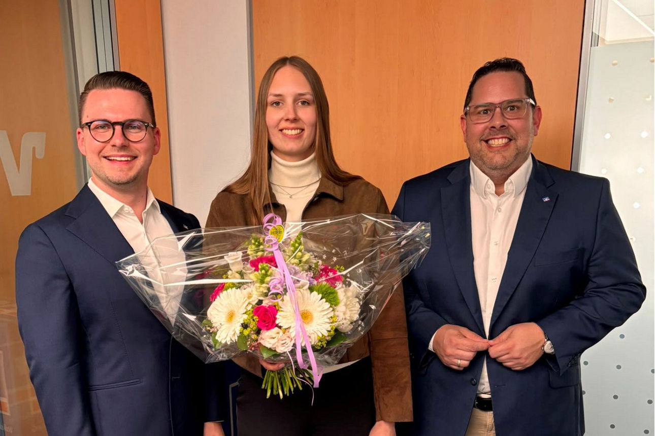 Zwei Männer in Anzügen flankieren eine junge Frau, die einen Blumenstrauß in der Hand hält
