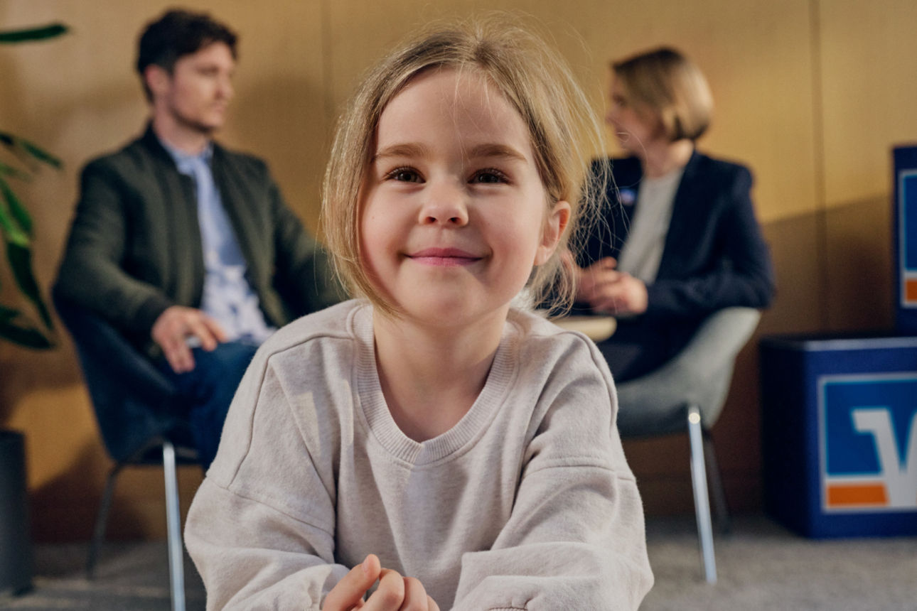 Ein kleines Mädchen sitzt an einem Tisch mit einem Malbuch und Stiften. Im Hintergrund wird ihr Vater gerade beraten.