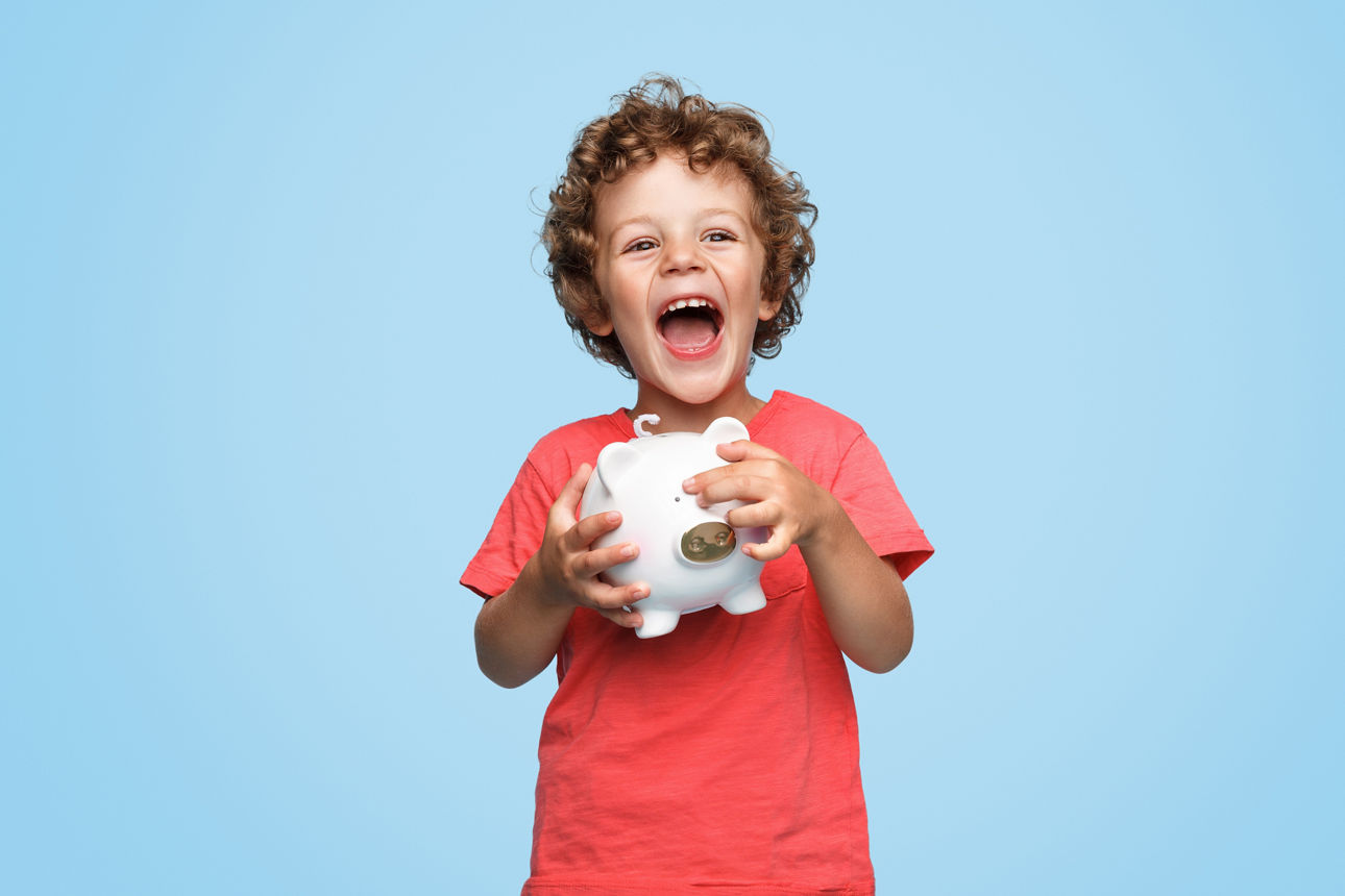 Cute little boy with piggy bank laughing and looking at camera while standing on blue background