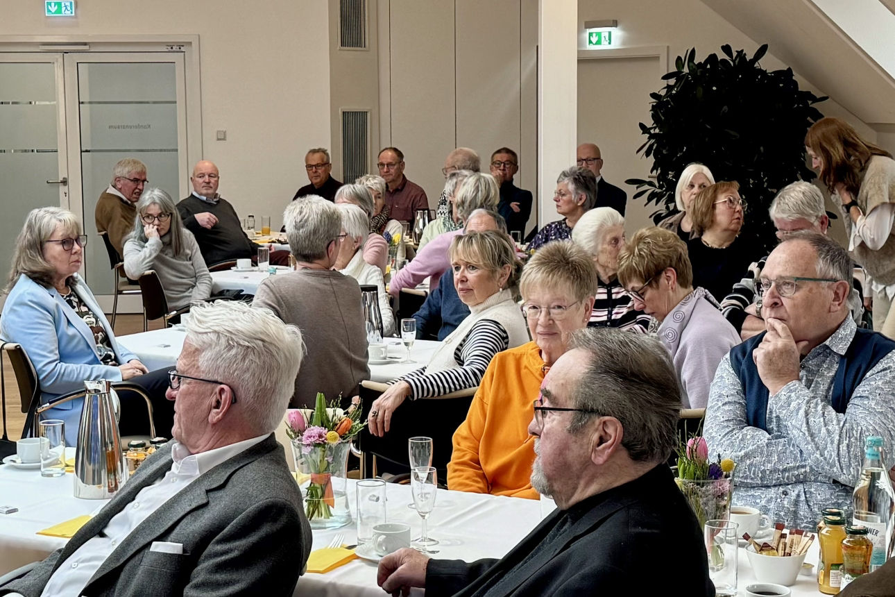 Mehrere ältere Frauen und Männer sitzen in einem hellen Konferenzraum an gedeckten Tischen mit Kaffee, Wasser und Blumenvasen und hören aufmerksam zu.