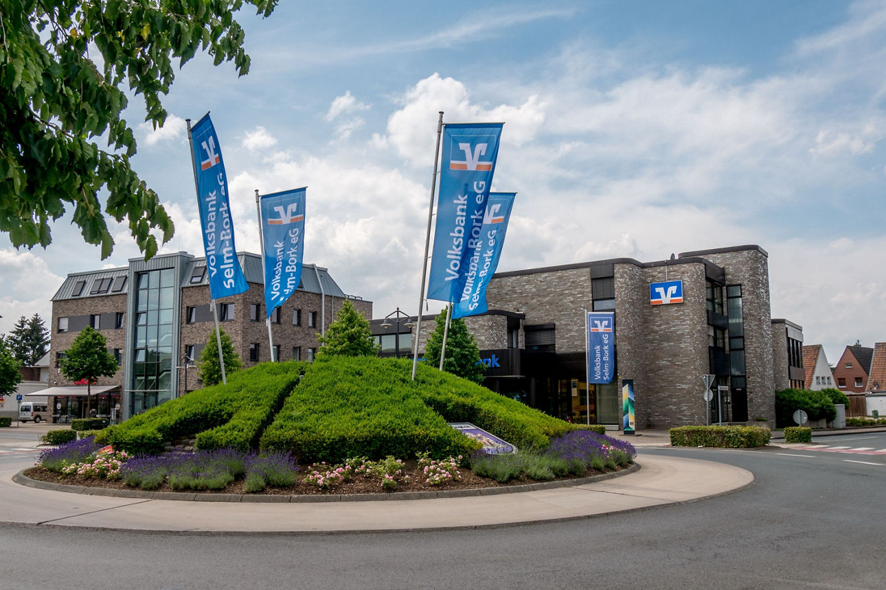 Eine Verkehrsinsel die bergrünt ist. Dahinter ist ein Volksbankgebäude zu sehen