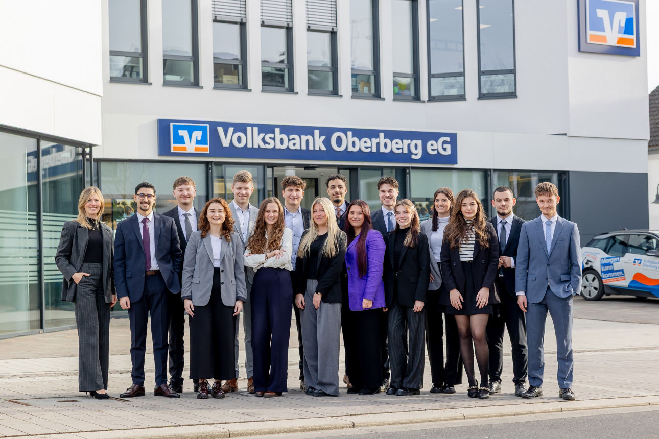 Gruppenfoto vor der Hauptgeschäftsstelle der Volksbank Oberberg in Wiehl. Links steht Ausbildungsleiterin Sarina Scherbaum, daneben die 15 neuen Bankkaufleute in Businesskleidung, im Hintergrund das Banklogo und die Fassadenfront.