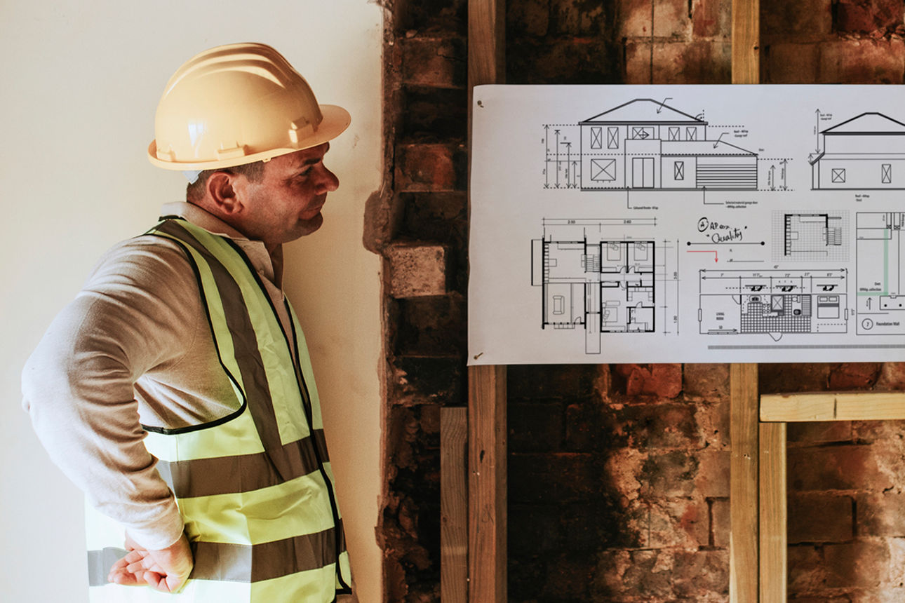 Zwei Handwerker begutachten Bauplan vor einer aufgestemmten Wand