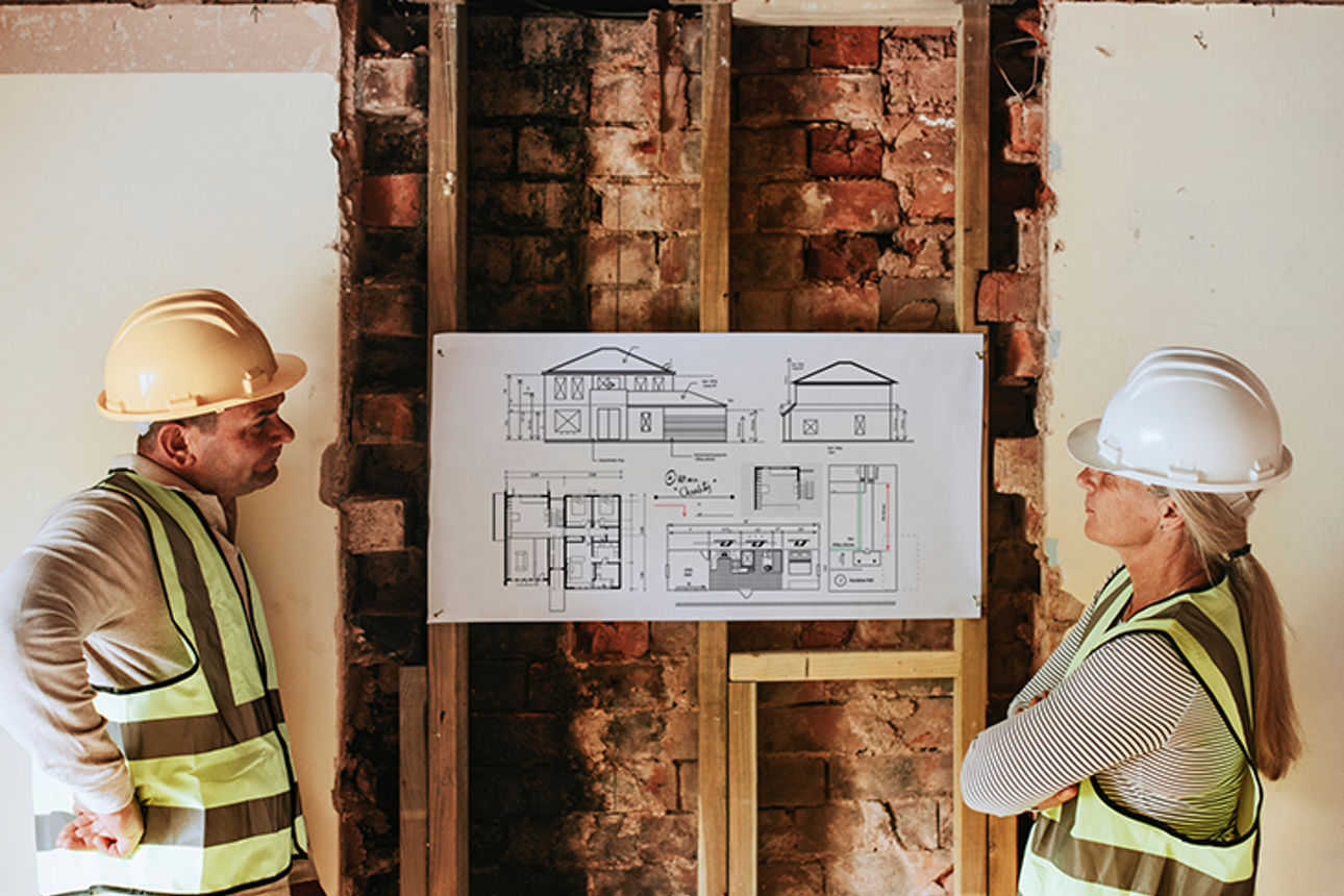 Zwei Handwerker begutachten Bauplan vor einer aufgestemmten Wand