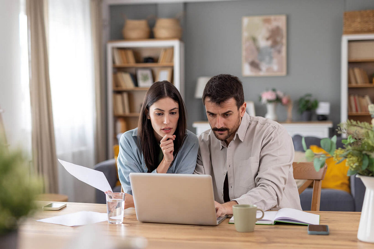 Junges Ehepaar sitzt am Schreibtisch vor dem Laptop und schaut auf den Bildschirm