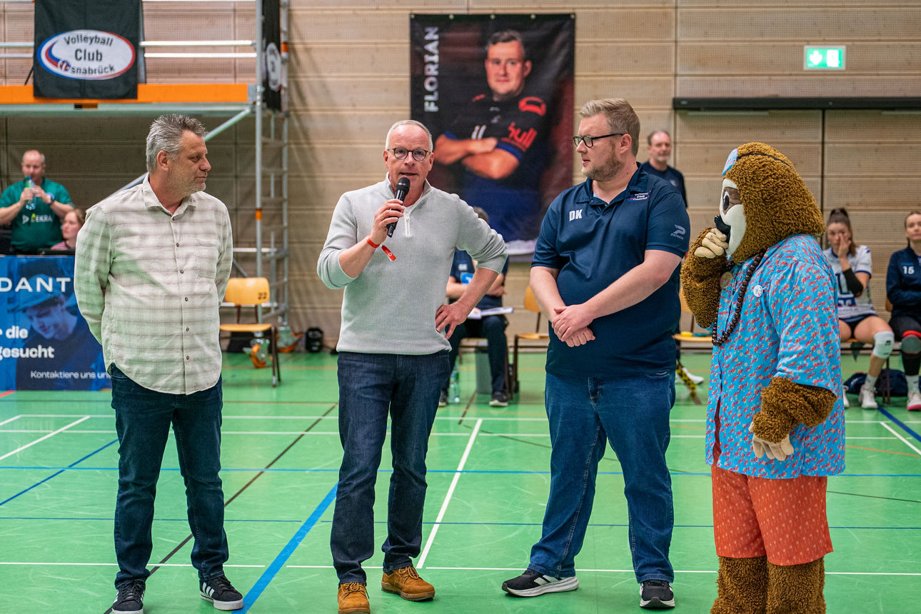 Das Maskottchenund drei Herren auf dem Spielfeld beim sprechen. 