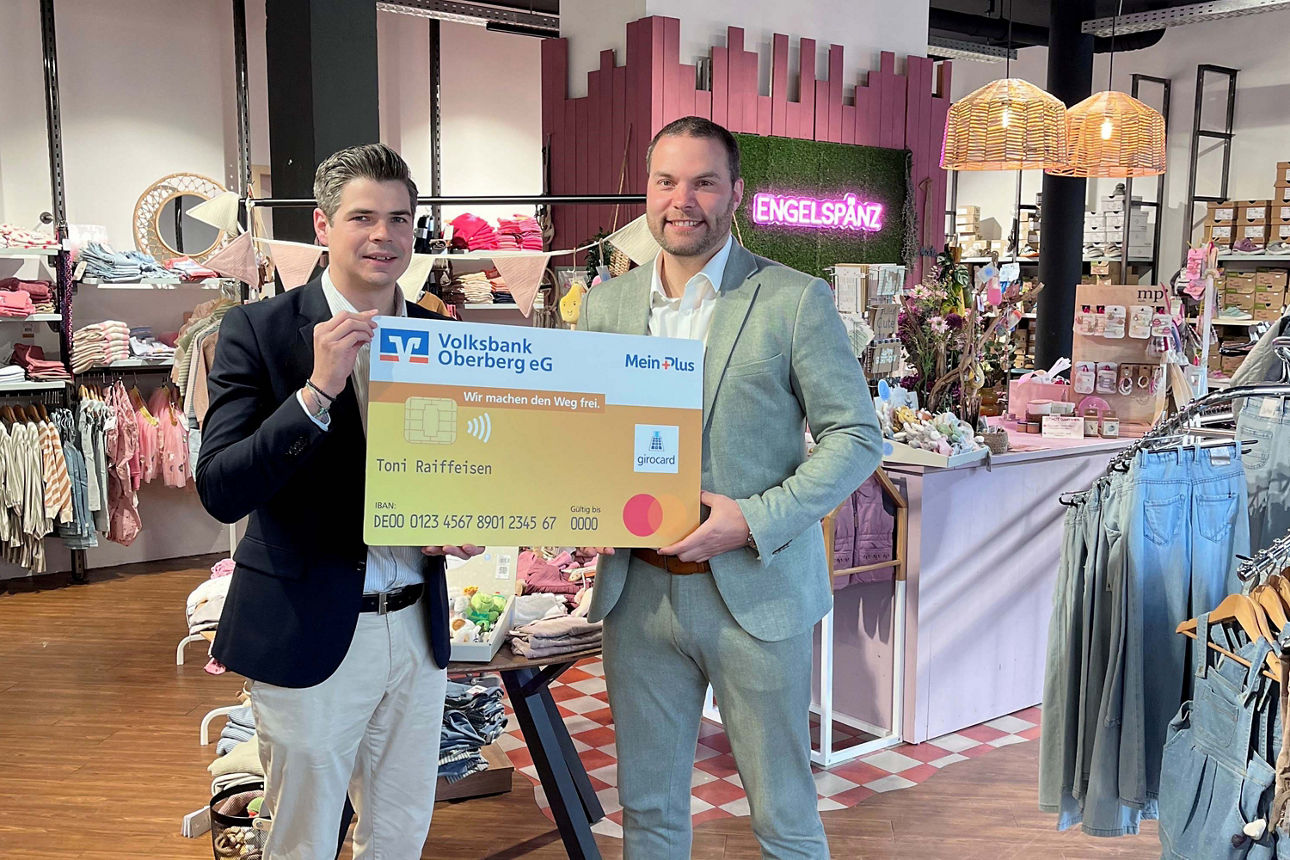 Zwei Männer lächeln in die Kamera und halten gemeinsam eine übergroße, symbolische Bankkarte der Volksbank Oberberg.