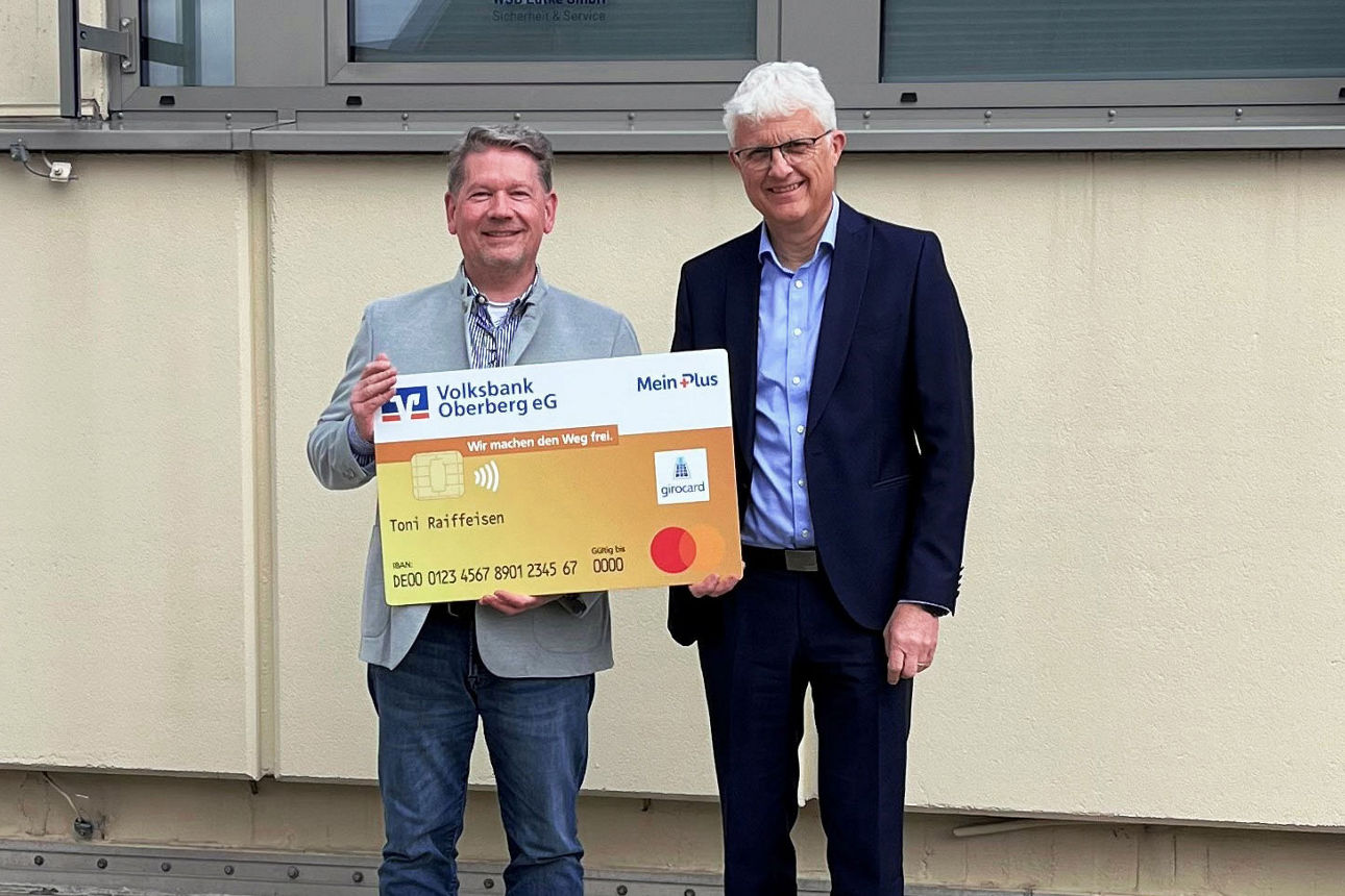 Zwei Männer lächeln in die Kamera und halten gemeinsam eine übergroße, symbolische Bankkarte der Volksbank Oberberg.