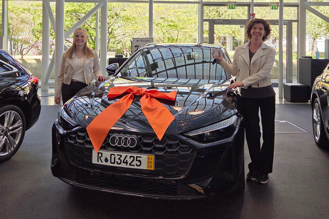 Zwei Damen stehen um ein schwarzes Auto herum. Das Auto hat eine orange Schleife auf der Motorhaube
