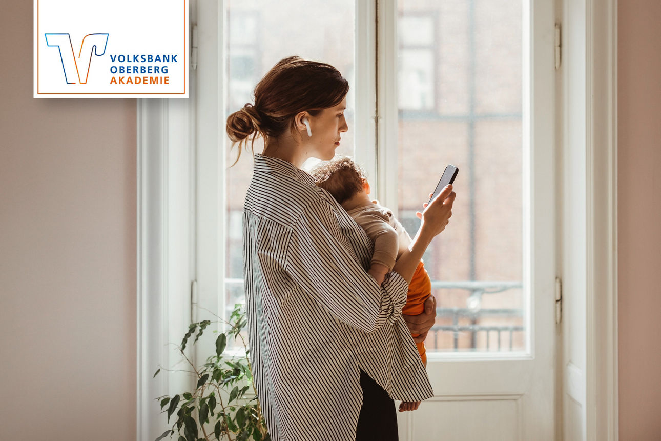 Frau hält ein Baby und schaut auf ihr Smartphone am Fenster; links oben Logo „Volksbank Oberberg Akademie“.