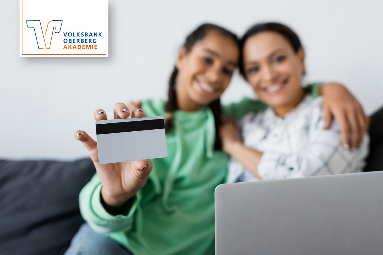 Zwei Personen zeigen eine Kreditkarte vor einem Laptop; links oben Logo „Volksbank Oberberg Akademie“.