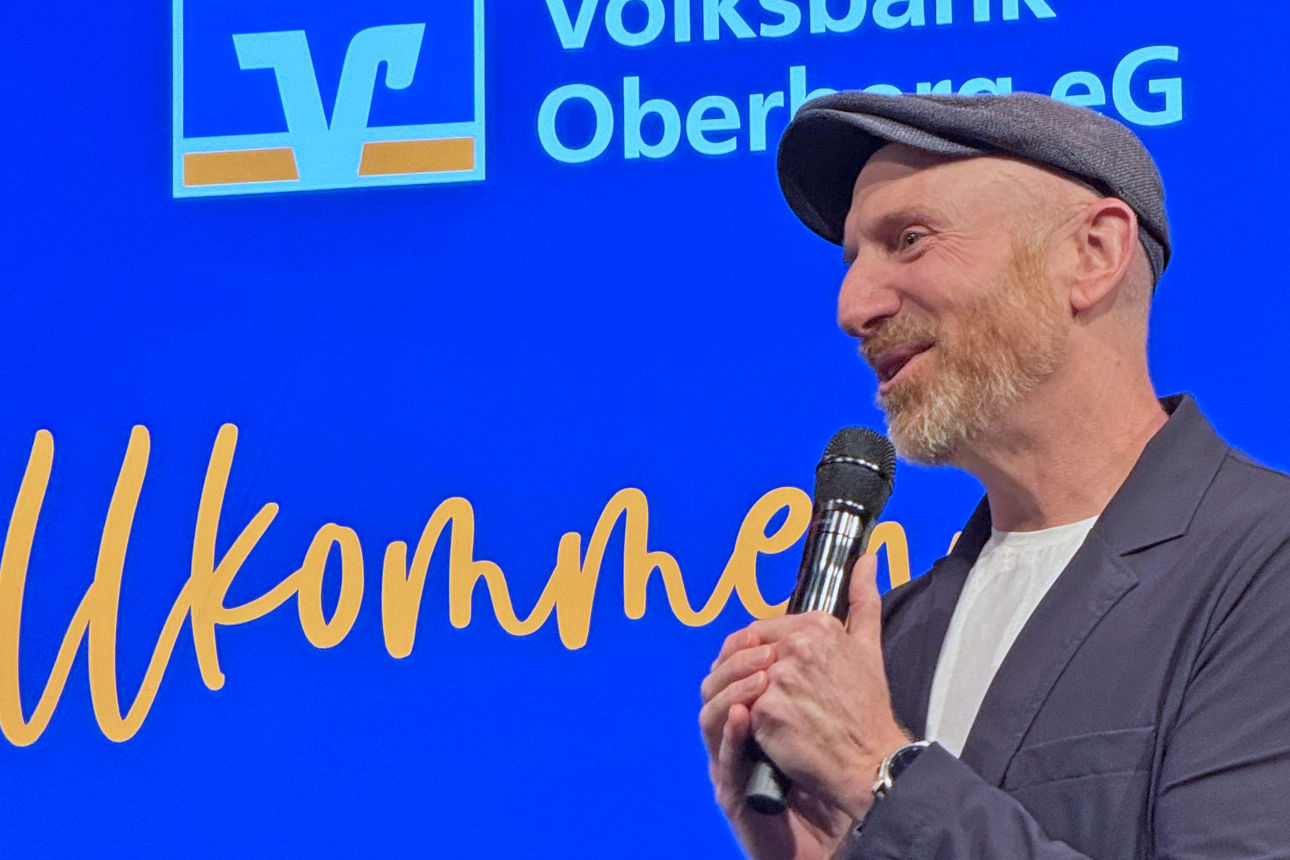 Martin Schopps steht auf der Bühne des Mitgliederabends der Volksbank Oberberg in Waldbröl und spricht mit Mikrofon vor blau beleuchtetem Bühnenhintergrund.