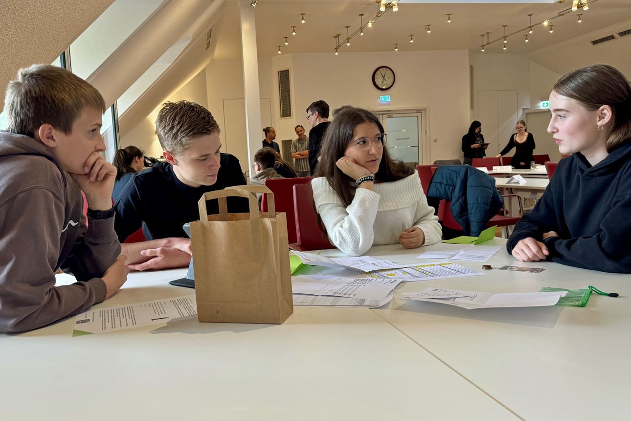 Mehrere Schülerinnen und Schüler arbeiten bei einem Workshop der Volksbank Oberberg in gemischten Kleingruppen gemeinsam an Unterlagen und Aufgaben des Mystery Games zur Buchhaltung.
