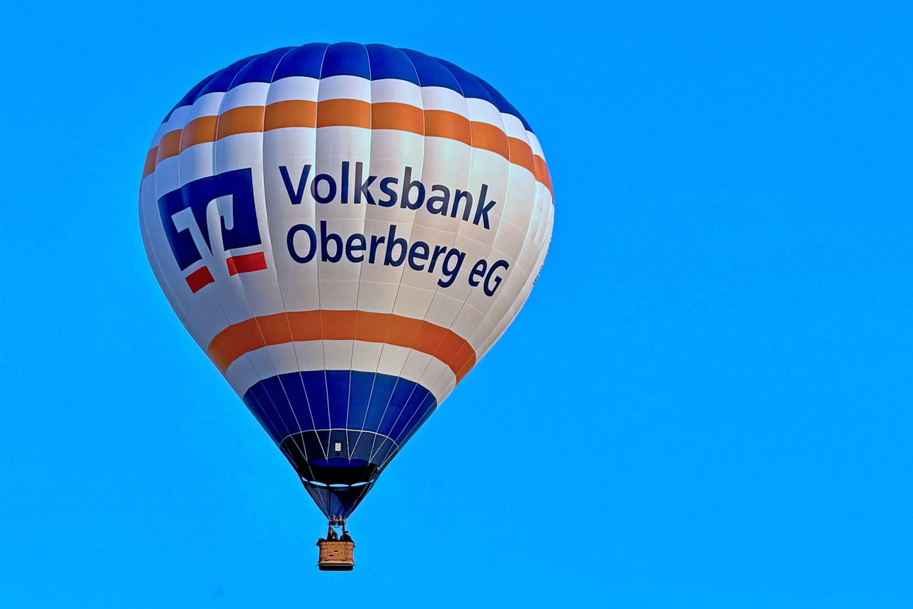 Der Heißluftballon der Volksbank Oberberg schwebt vor blauem Himmel. Auf der weißen Ballonhülle sind das Logo und der Schriftzug der Volksbank Oberberg zu sehen.