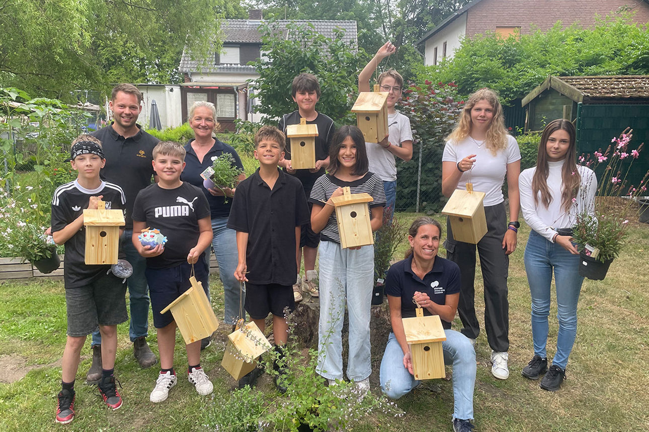 Eine Gruppe von Schülern und Erwachsenen hält Vogel-Nistkästen und Blumen in den Händen.