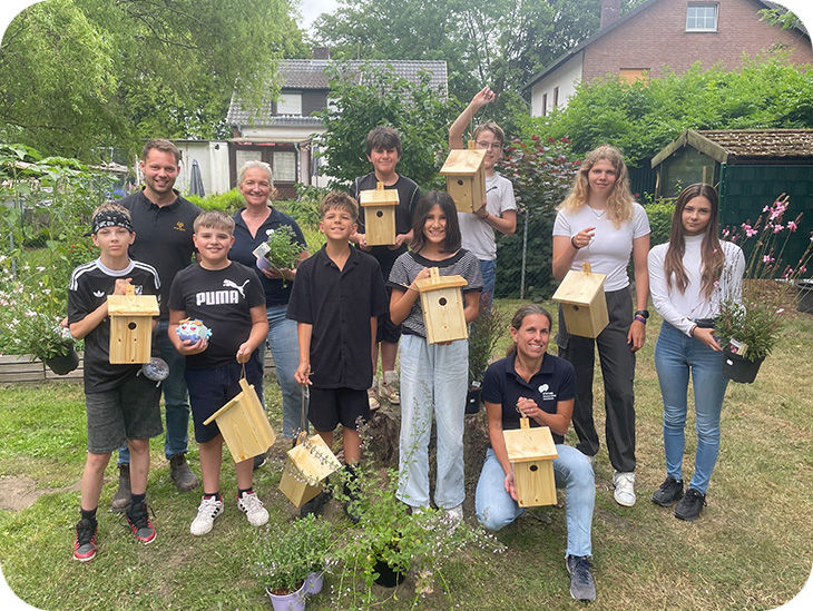 Eine Gruppe von Schülern und Erwachsenen hält Vogel-Nistkästen und Blumen in den Händen.