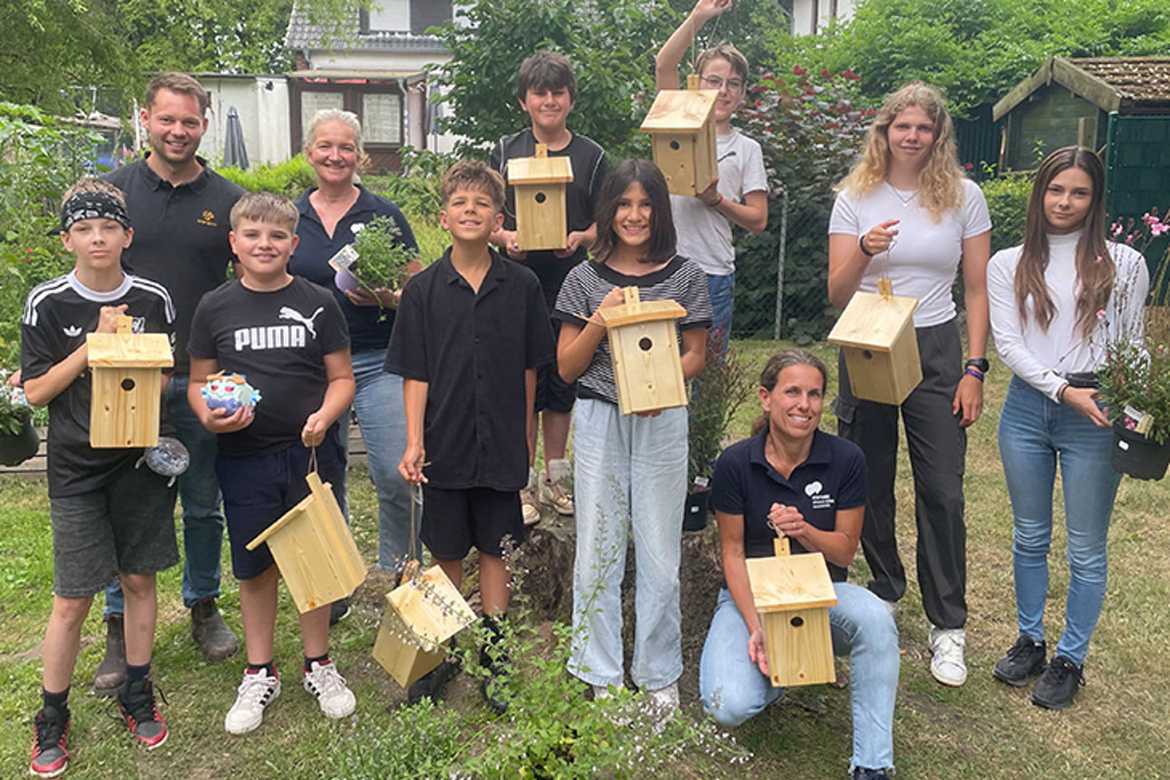 Eine Gruppe von Schülern und Erwachsenen hält Vogel-Nistkästen und Blumen in den Händen