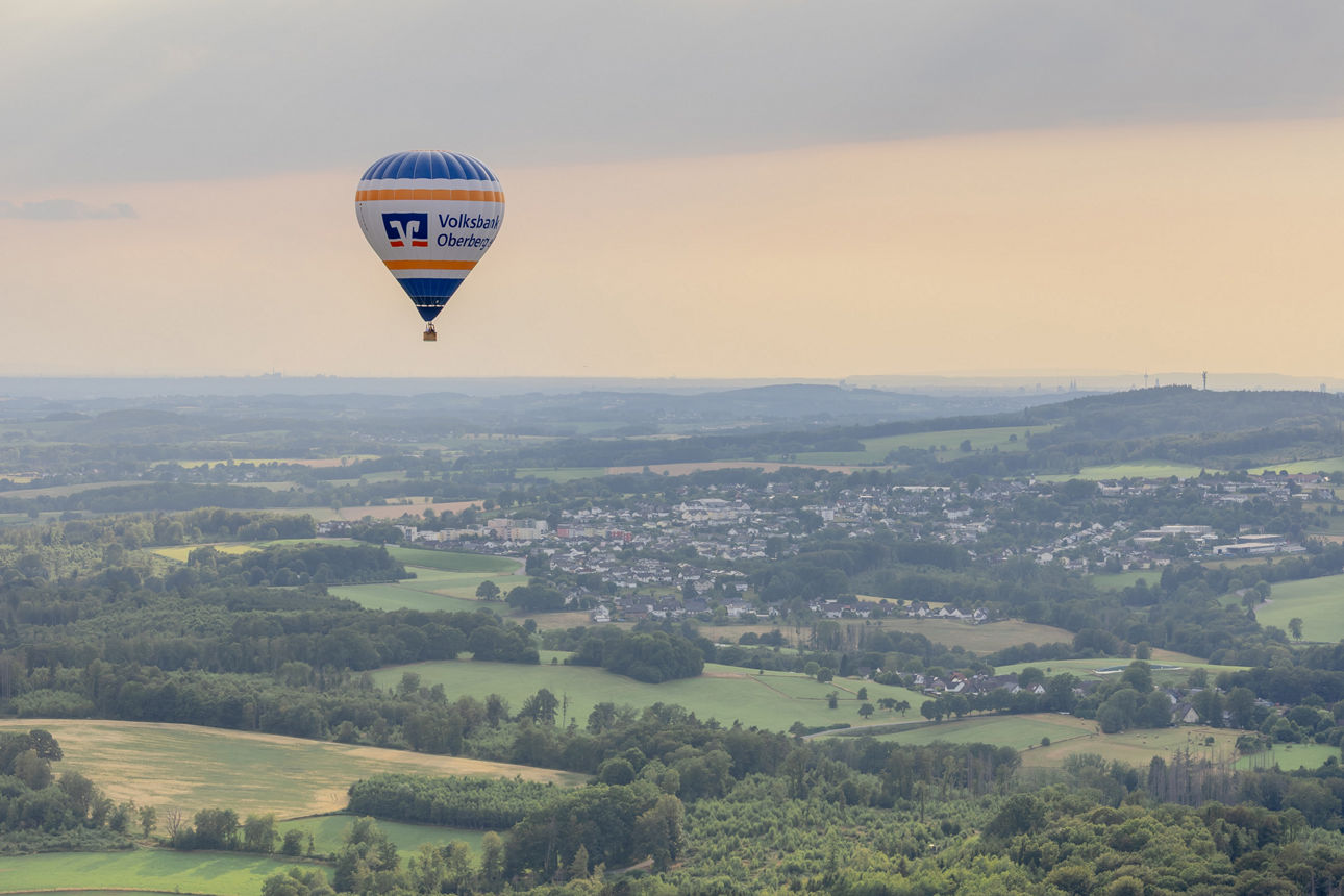 Der Heißluftballon D-OVOB der Volksbank Oberberg fliegt über die hügelige Landschaft des Oberbergischen Kreises. Im Hintergrund sind Ortschaften, Felder und Wälder unter einem Abendhimmel mit aufziehenden Regenwolken zu sehen.