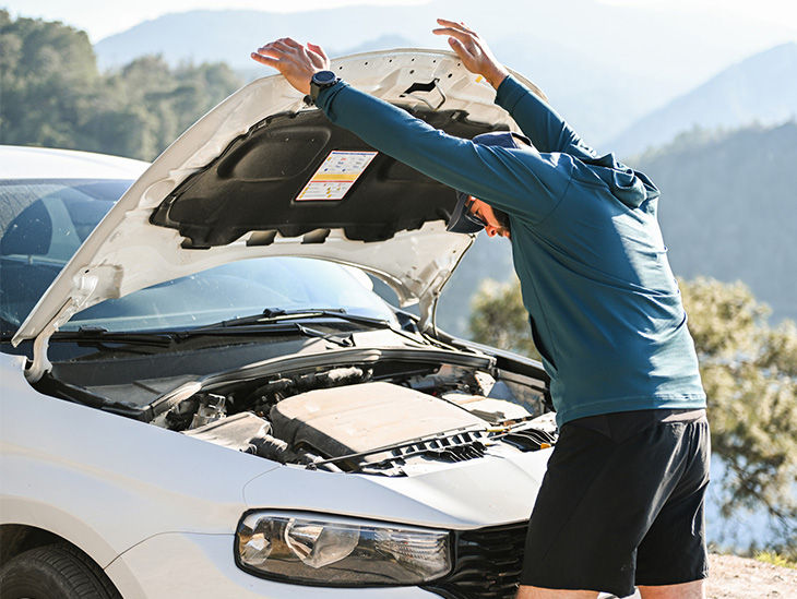Mann vor Urlaubskulisse steht vor seinem kaputten Auto und öffnet die Motorhaube. 