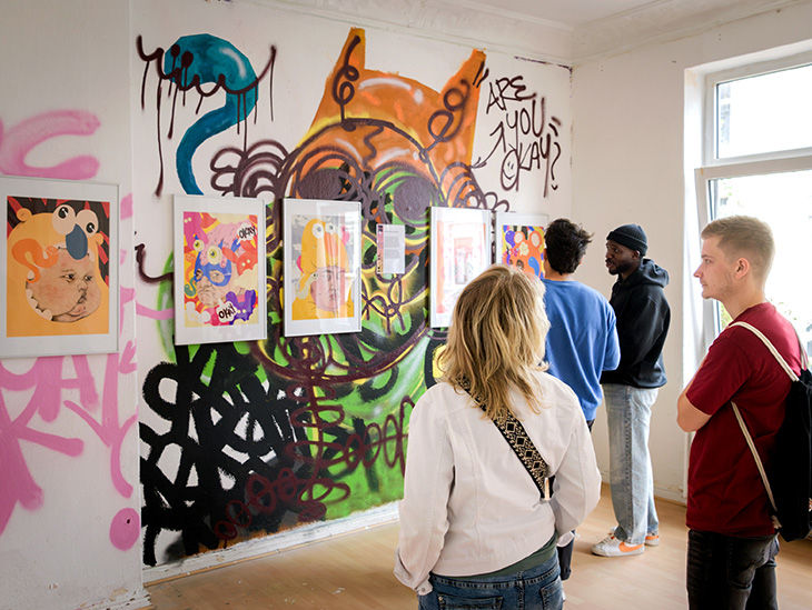Besucherinnen und Besucher betrachten Bilder. Die Bilder hängen an einer mit Graffiti besprayten Wand.