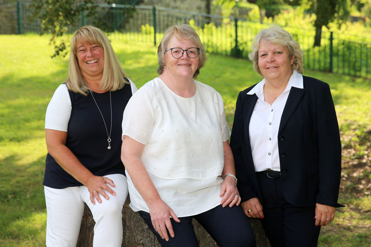 Claudia Koschwitz, Anette Nolte und Anja Ufer sitzen und stehen lächelnd auf einem Baumstamm im Wiehlpark in der Nähe der Hauptstelle Wiehl.