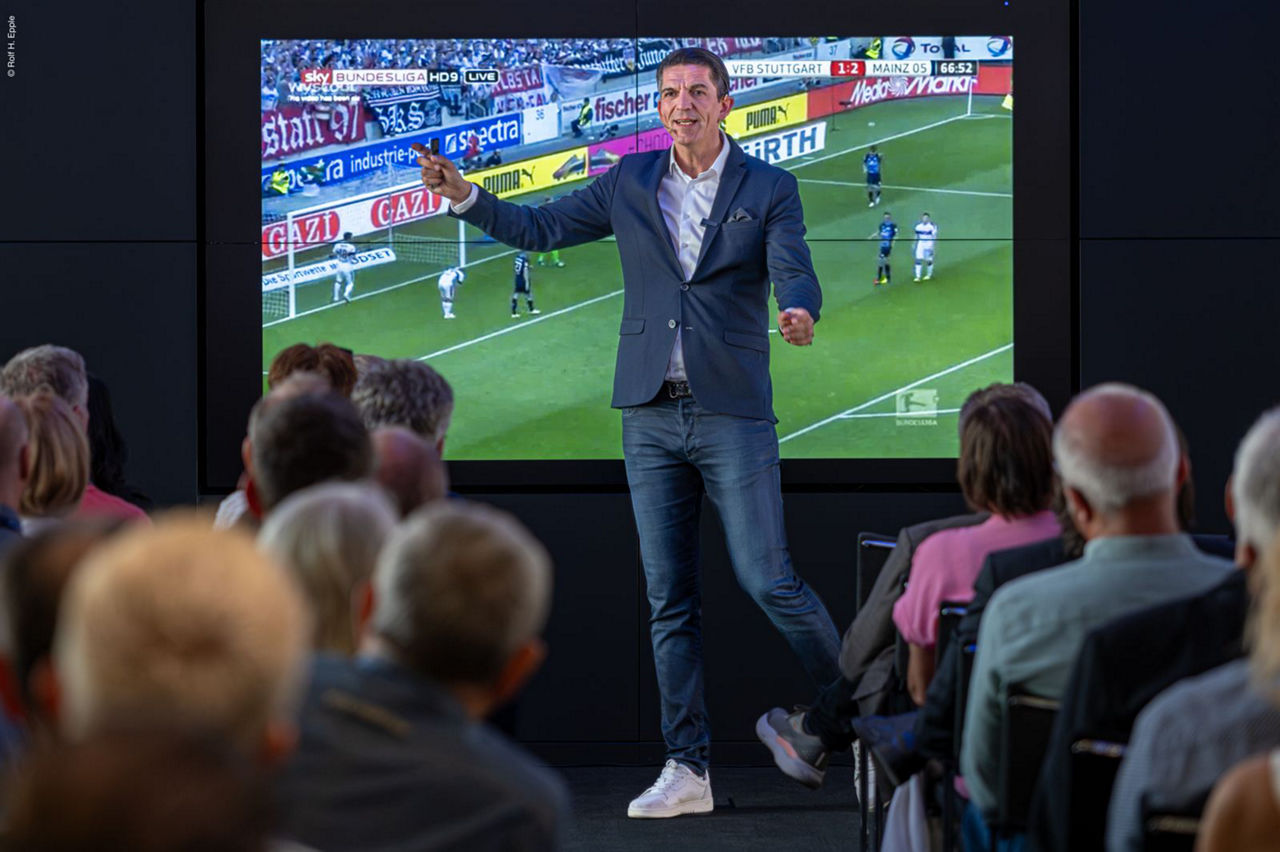 Deniz Aytekin steht vor dem Plenum während im Hintergrund eine Szene aus der Bundesliga erklärt wird.