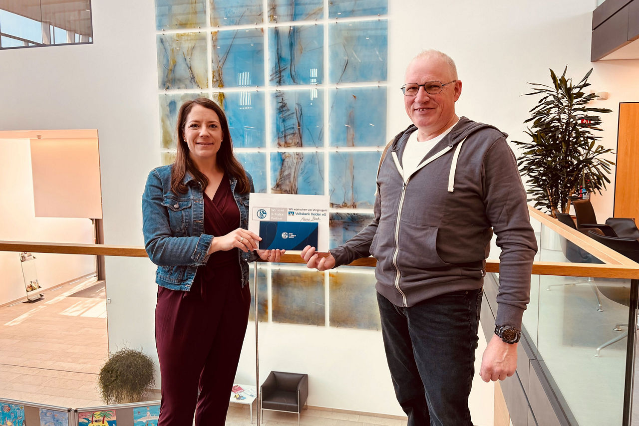 Ein Mann und eine Frau stehen in der Lobby eines modernen Gebäudes und halten ein Zertifikat mit Markenlogo in der Hand.