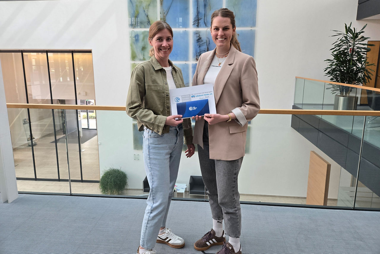 Zwei Frauen stehen in der Galerie eines modernen Gebäudes und halten ein Zertifikat mit Markenlogo in der Hand.