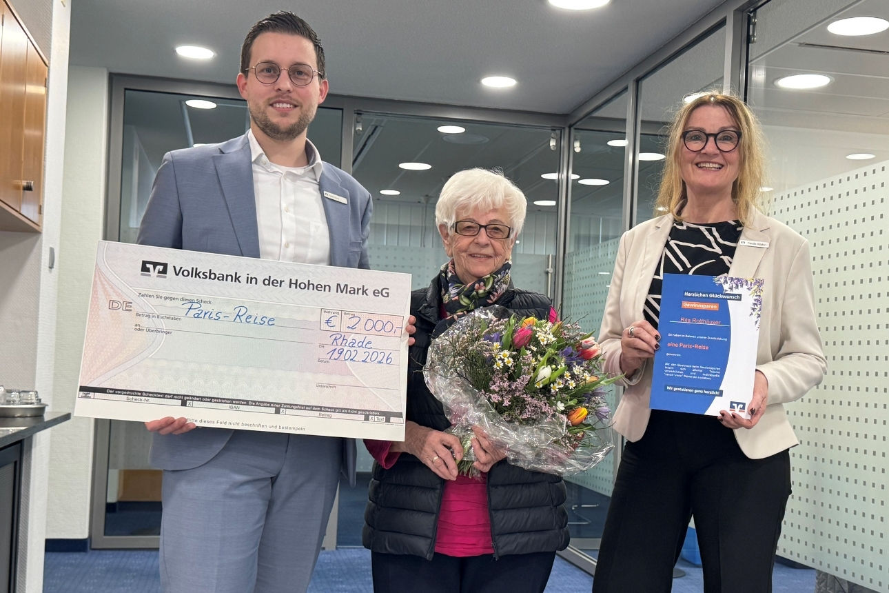 Drei Personen stehen in einem Bankgebäude. Eine Person hält einen großen symbolischen Scheck der Volksbank in der Hohen Mark eG über 2.000 Euro mit dem Vermerk „Paris-Reise“, eine weitere Person hält einen Blumenstrauß. Auf einer Urkunde wird ein Gewinn beim Gewinnsparen für eine Paris-Reise erwähnt.