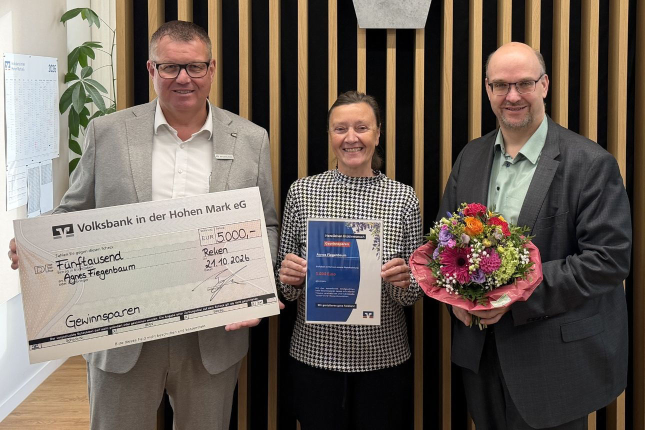Drei Personen stehen in einer Bankfiliale: Der Kundenberater hält einen symbolischen Scheck über 5.000 Euro aus dem Gewinnsparen, die Gewinnerin eine Urkunde und ein Vorstandsmitglied der Bank überreicht einen Blumenstrauß. 