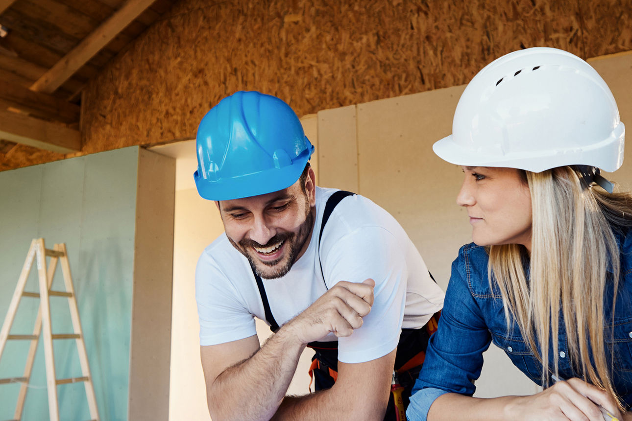Mann mit blauem und Frau mit weißem Helm unterhalten sich auf Baustelle.