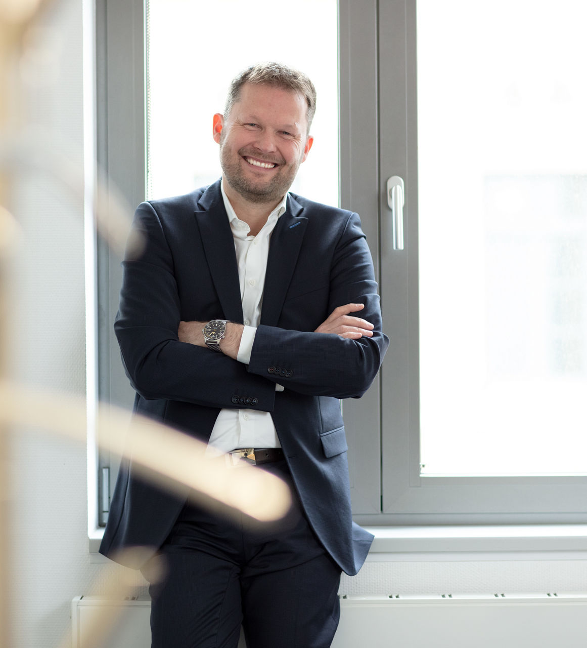 Ein Mann im Anzug, der lächelnd und mit verschränkten Armen vor einem Fenster steht
