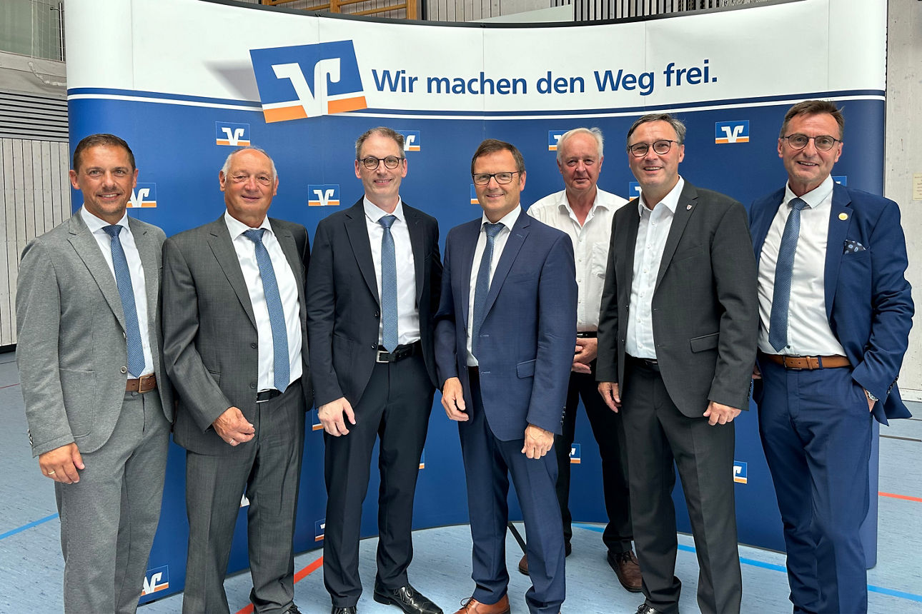 Eine Gruppe von sieben Männern in Geschäftskleidung posiert vor einer Kulisse mit dem Volksbank-Logo für ein Foto.