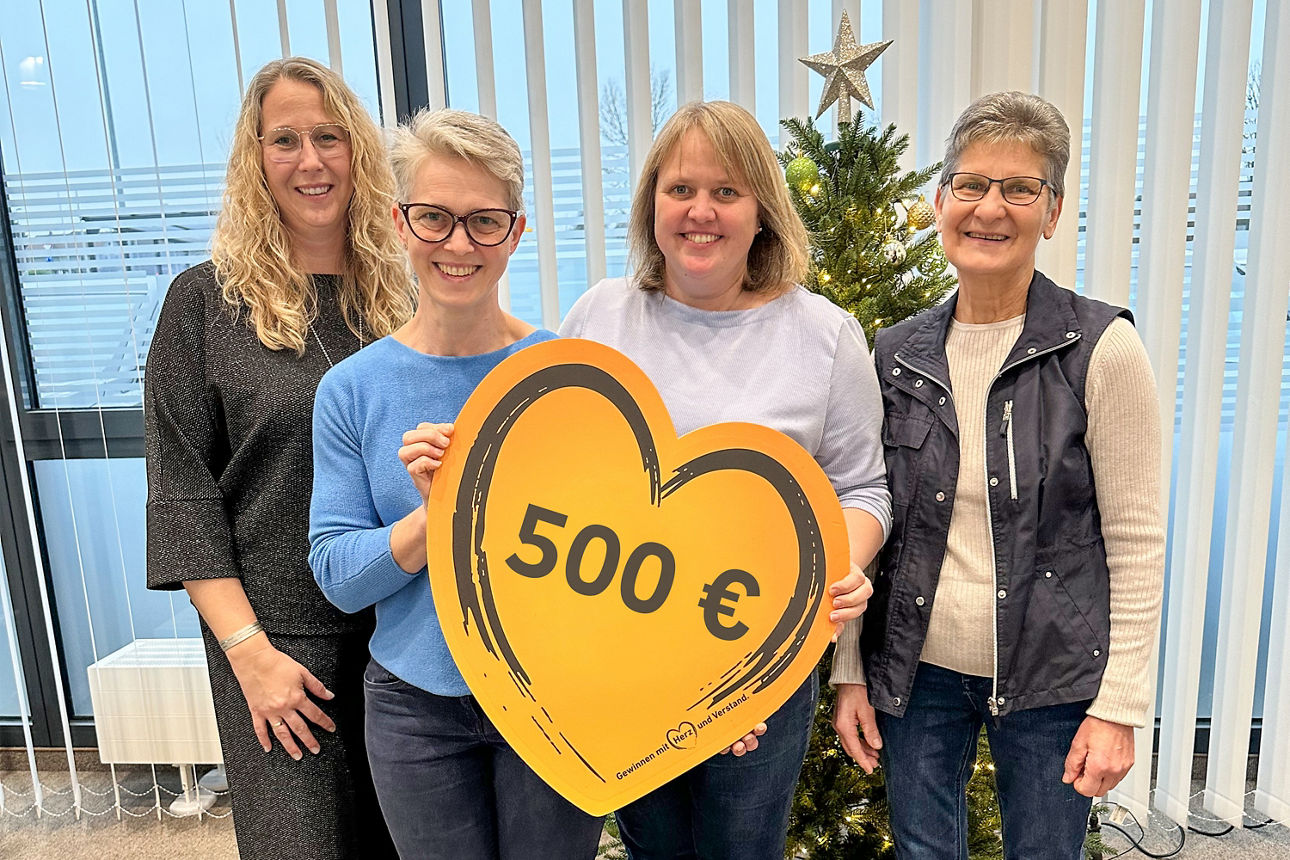 Vier Frauen posieren in einem Innenraum mit einem großen, herzförmigen Schild, auf dem ein Wert von 500 € angegeben ist.