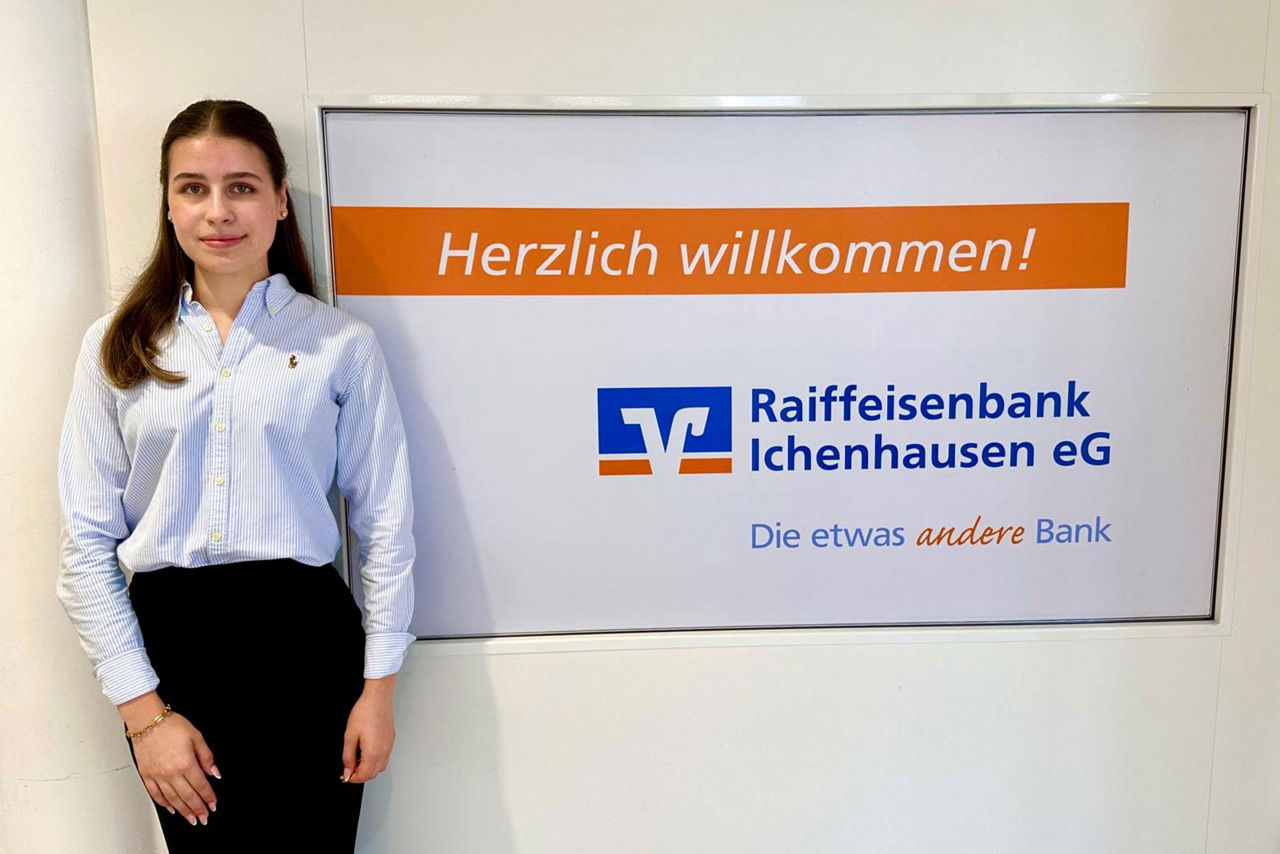 Elisa Joas vor einem Schild der Raiffeisenbank Ichenhausen eG, auf dem "Herzlich willkommen!" steht