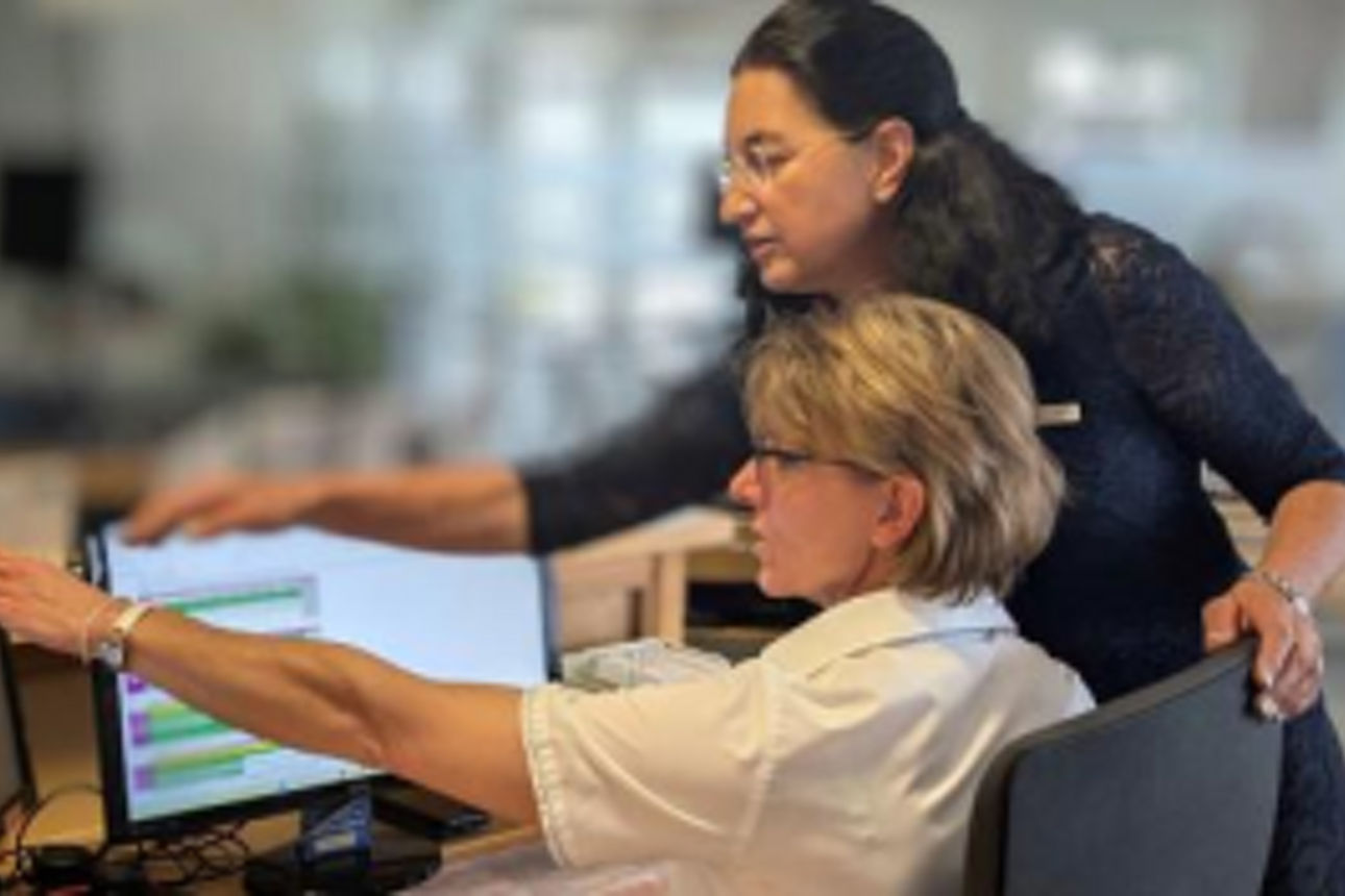 Zwei Personen im Büro, eine am Computer mit Diagrammen, die andere steht beratend daneben