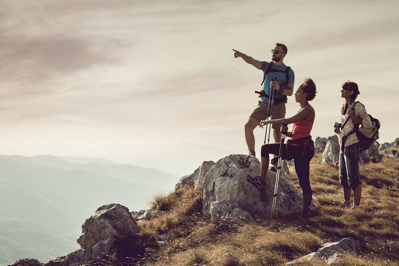 Drei Wanderer auf einem Berg, einer der drei deutet mit dem Arm auf etwas in der Ferne