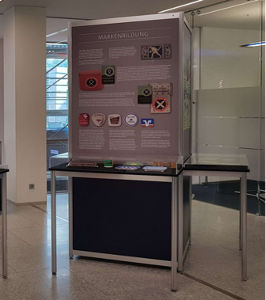 Ausstellungstafel mit Texten, Logos und Objekten auf einem Tisch in modernem Innenraum