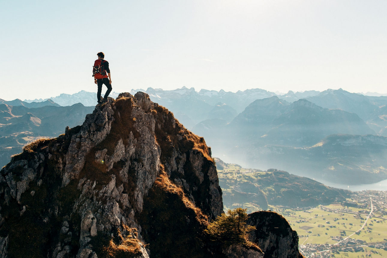 Eine Person steht mit Rucksack auf einem felsigen Gipfel, im Hintergrund ein weites Alpenpanorama.