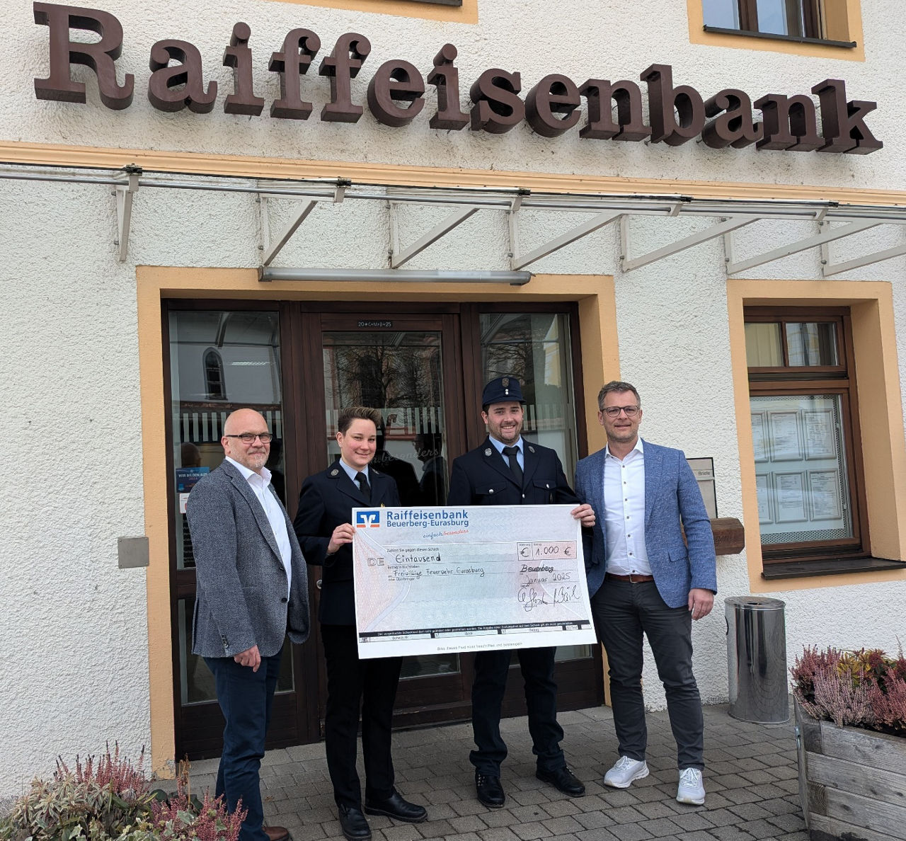 Vier Personen vor Raiffeisenbank-Gebäude mit großem Spendenscheck in den Händen