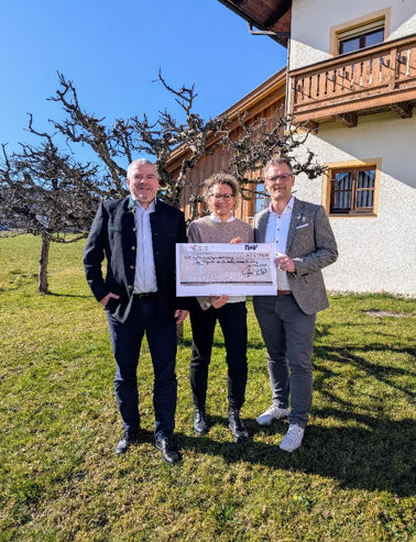 Drei Personen mit großem Scheck über 2.000 € vor Haus mit Balkon und Bäumen im Hintergrund