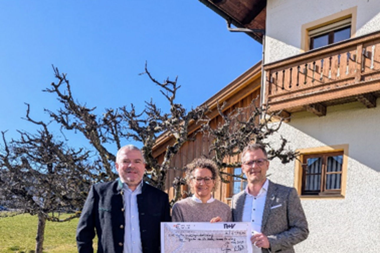 Drei Personen mit großem Scheck über 2.000 € vor Haus mit Balkon und Bäumen im Hintergrund